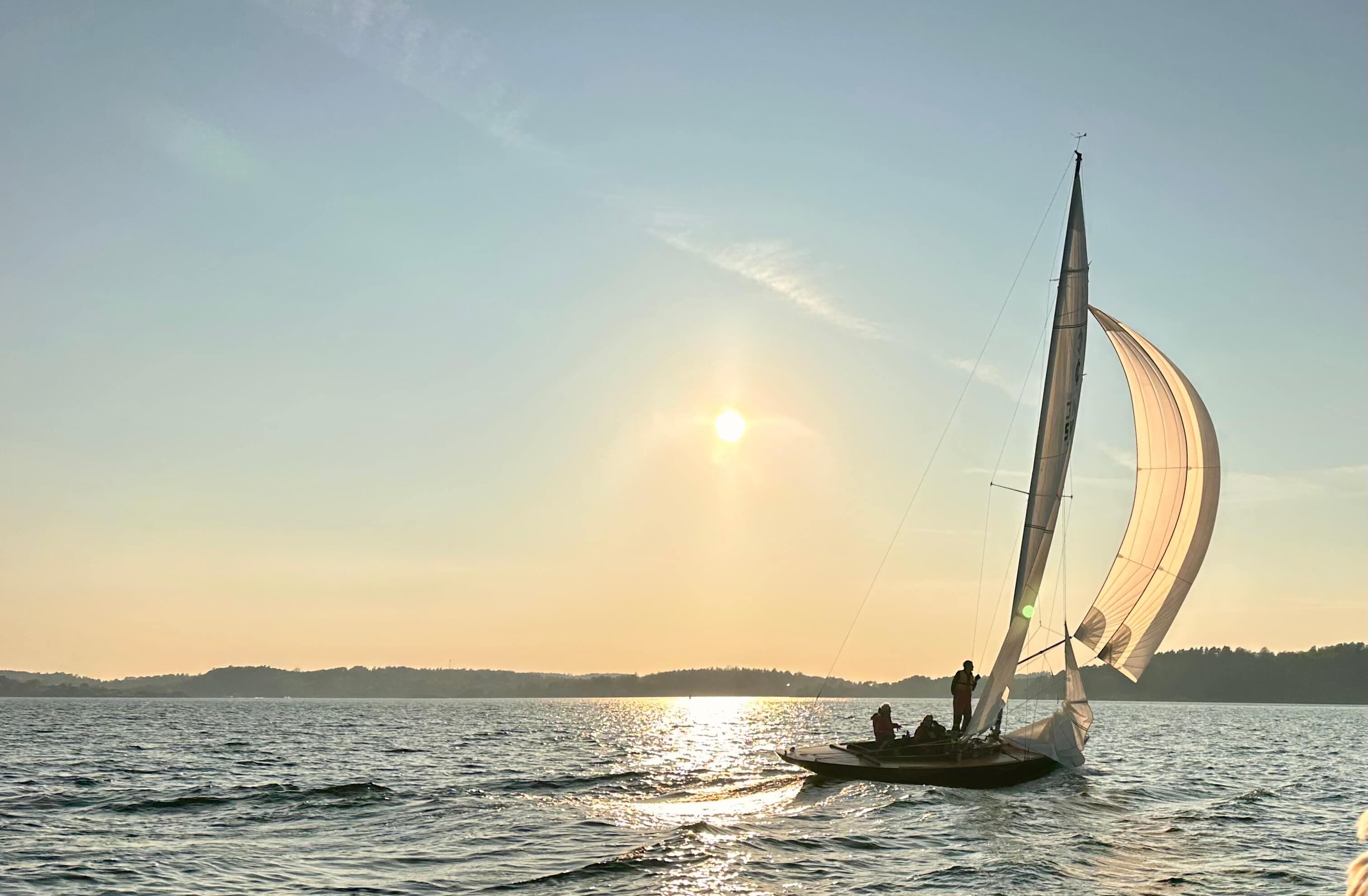 An International One Design under spinnaker in low evening sun, the mainland shore across the fjord beyond